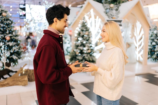 Seitenansicht zu einem fröhlichen jungen Mann, der der liebevollen blonden Freundin, die am Weihnachtsabend in der Halle des Einkaufszentrums steht, eine Geschenkbox mit Geschenk überreicht