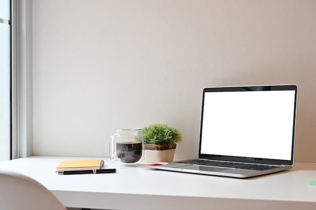 Seitenansichtarbeitsplatz-Computerlaptop, Kaffee, Notizbuch mit Stift auf weißer Tabelle.