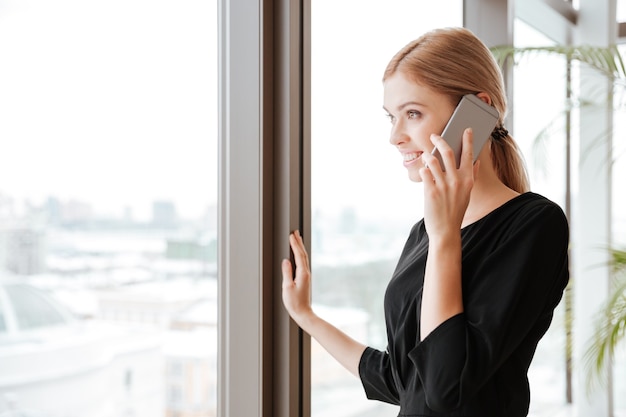 Seitenansichtbild einer jungen Arbeiterin, die im Büro steht, während sie auf das Fenster schaut und mit ihrem Telefon spricht.