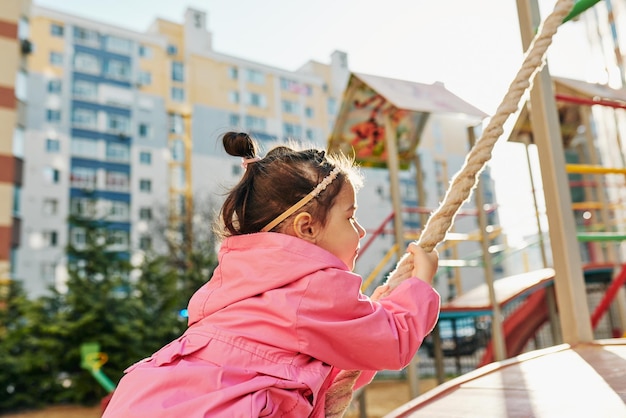 Seitenansichtbild eines glücklichen hübschen kleinen Mädchens, das auf einem Spielplatz im Freien an einem Seil klettert Niedliches Kleinkindmädchen im Vorschulalter, das außerhalb des Bildungs- und Aktivitätskonzepts der Kindheit spielt