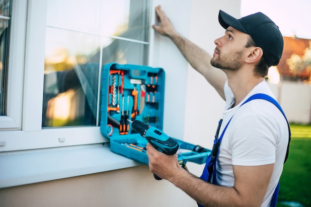 Seitenansichtfoto eines jungen Arbeiters in Uniform, während er im Freien Fenster installiert oder repariert