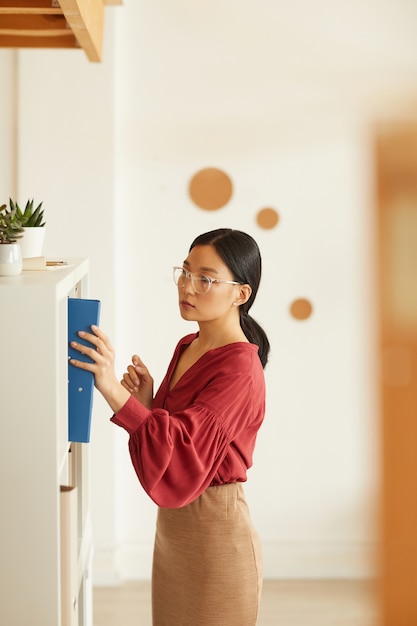 Seitenansichtporträt der eleganten asiatischen Geschäftsfrau, die Binder zum Bücherregal gegen weiße Wand im Büro, Kopierraum setzt