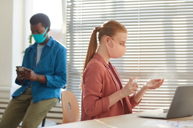 Seitenansichtporträt der jungen Frau, die Gesichtsmaske trägt, die Hände am Arbeitsplatz im Postpandemiebüro desinfiziert