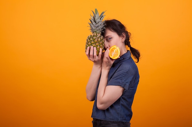 Seitenansichtporträt der schönen jungen Frau, die leckere Ananas und Orange im Studio über gelbem Hintergrund hält. Sommer leckere Früchte.