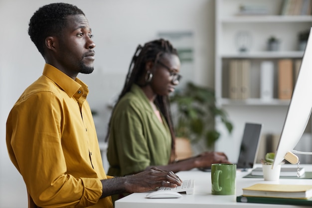 Seitenansichtporträt des jungen afroamerikanischen Mannes, der PC im modernen Büro verwendet und Computerbildschirm, Kopienraum betrachtet