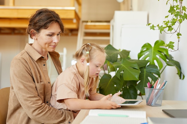 Seitenansichtporträt des niedlichen Mädchens mit Down-Syndrom unter Verwendung der digitalen Tablette mit Mutter beim Lernen zu Hause, Kopienraum