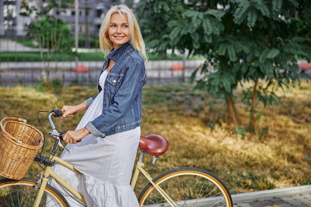 Seitenansichtporträt einer attraktiven blonden kaukasischen Frau in einem stilvollen weißen langen Kleid, das ihre Hände am Lenker des Citybikes mit Korb hält holding