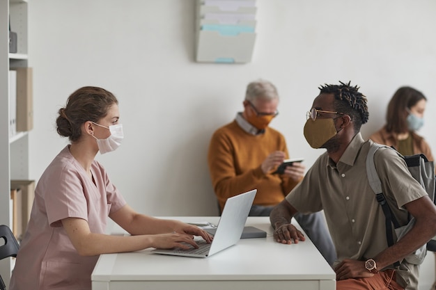 Seitenansichtporträt einer Krankenschwester, die Patienten registriert, die in der Arztpraxis in Reihe warten, alle tragen Masken, Kopierraum