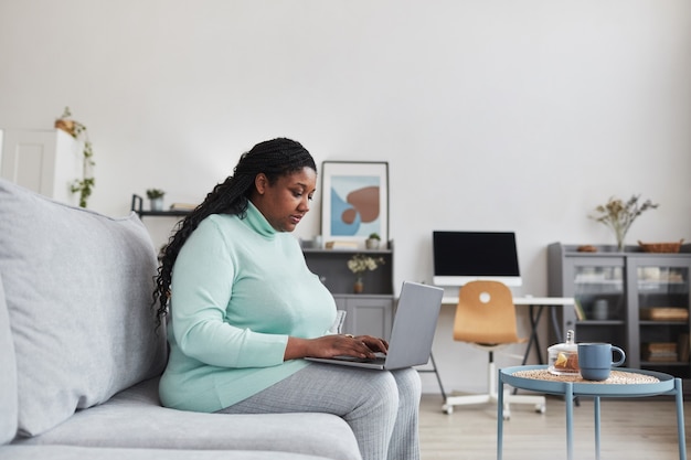 Seitenansichtporträt einer kurvigen Afroamerikanerin, die Laptop benutzt, während sie im Internet auf der Couch in minimalem Innenraum sitzt, Kopienraum