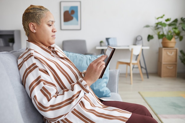 Seitenansichtporträt einer modernen gemischtrassigen Frau, die ein digitales Tablet hält und Online-Dienste nutzt, während sie zu Hause auf dem Sofa sitzt, Platz kopieren