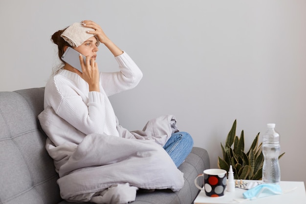 Foto seitenansichtporträt einer ungesunden dunkelhaarigen frau, die einen weißen pullover trägt, der auf husten sitzt, mit dem arzt spricht, die hand mit einem handtuch auf der stirn hält, unter kopfschmerzen und hoher temperatur leidet.