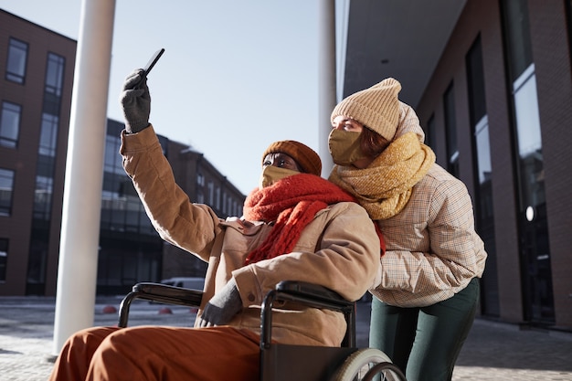 Seitenansichtporträt eines afroamerikanischen Mannes, der Rollstuhl benutzt und Maske trägt, während er ein Selfie-Foto mit junger Frau macht, die in der Stadt hilft, Raum kopieren