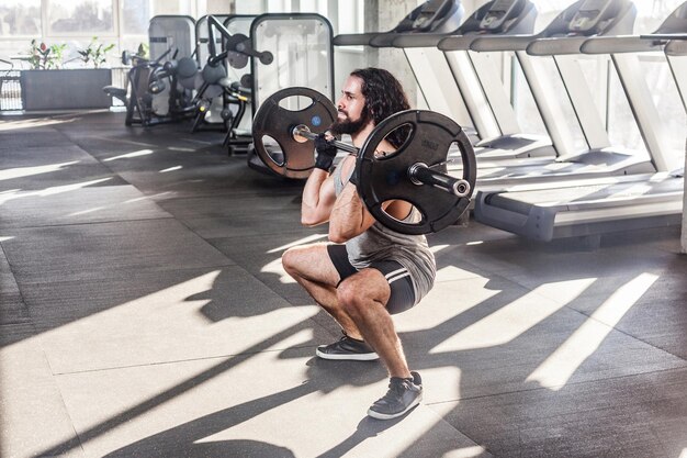 Seitenansichtporträt eines jungen erwachsenen Bodybuilders mit langen lockigen Haaren trainieren allein im Fitnessstudio in der Hocke und bereiten sich auf das Heben der Langhantel vor, die Übungen für gesundes Innenkonzept der Beinmuskeln macht