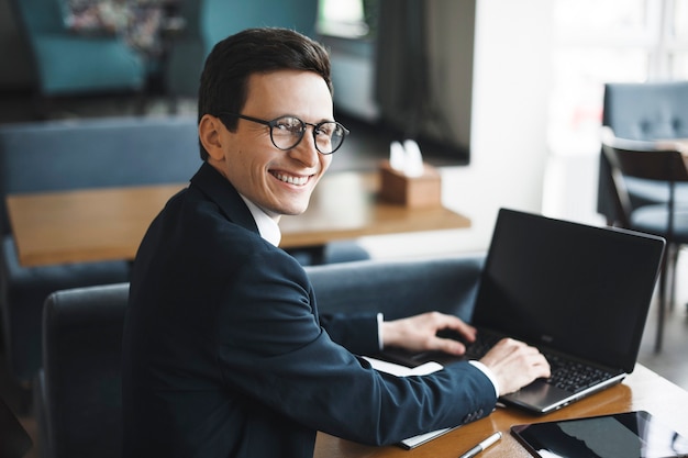 Seitenansichtporträt eines stilvollen erwachsenen kaukasischen Managers, der Anzug und Brille trägt, die über seine Schulter lächeln, während am Laptop im Café arbeiten.