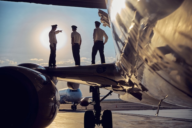 Seitenansichtporträt von drei nicht erkennbaren Personen, die auf den Flügel eines großen Passagierflugzeugs stehen und in den sonnigen Himmel schauen