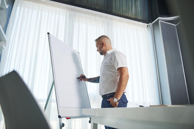 Seitenporträt eines kaukasischen Mannes mittleren Alters in weißem Freizeithemd und tätowierten Armen, der einen Geschäftsplan auf Flipchart schreibt