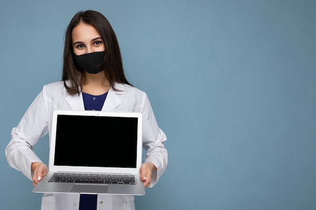 Seitenprofilaufnahme der jungen Brunetfrau, die medizinischen weißen Mantel und schwarze Maske hält, die Laptop hält und Kamera lokalisiert auf blauer Wand betrachtet