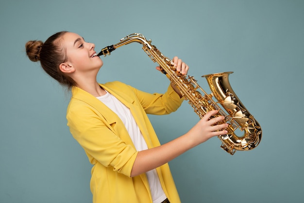 Seitenprofilfoto einer hübschen, positiv lächelnden, brünetten Teenagerin, die eine trendige gelbe Jacke trägt, die isoliert über einer blauen Hintergrundwand steht und Saxophon spielt, das zur Seite schaut