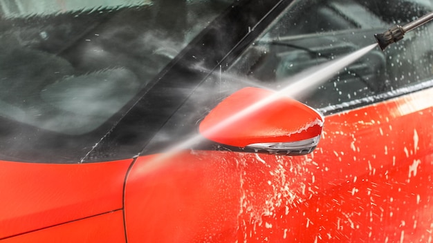 Seitenspiegel des roten Autos in Selbstbedienungswaschanlage gewaschen, Strahlwasserstrahl sprüht aus Schlauchdüse.