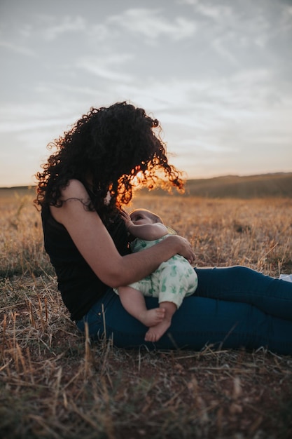 Seitliche Ansicht der Mutter, die das Baby stillt, während sie während des Sonnenuntergangs auf dem Feld gegen den Himmel sitzt