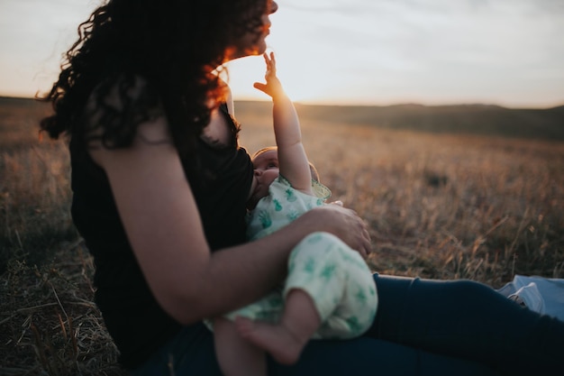 Seitliche Ansicht der Mutter, die das Baby stillt, während sie während des Sonnenuntergangs auf dem Feld gegen den Himmel sitzt