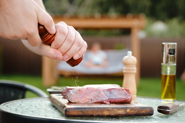 Seitliche Ansicht eines unerkennbaren Mannes, der Pfeffer zum rohen Rindfleischsteak hinzufügt