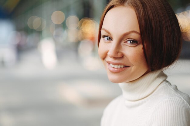 Seitliche Aufnahme einer angenehm aussehenden Frau mit geschminktem zartem Lächeln in weißem Rollkragenpullover steht vor verschwommenem Hintergrund mit Kopierfläche für Ihre Werbung hat gesunde Haut
