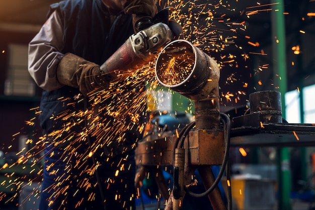 Seitliche Nahaufnahme des professionellen fokussierten fleißigen Mannes in der Uniform, die an der Metallrohrskulptur mit einem elektrischen Schleifer arbeitet, während Funken in der industriellen Stoffwerkstatt fliegen.