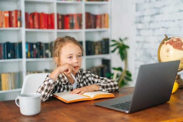 Seitliche Nahaufnahme eines ernsthaften kaukasischen Schulmädchens, das Hausaufgaben macht, das zu Hause mit Laptop und Notizbuch aus Papier am Tisch sitzt Adorable Grundschülerin Schulmädchen elearning online mit Computer-App