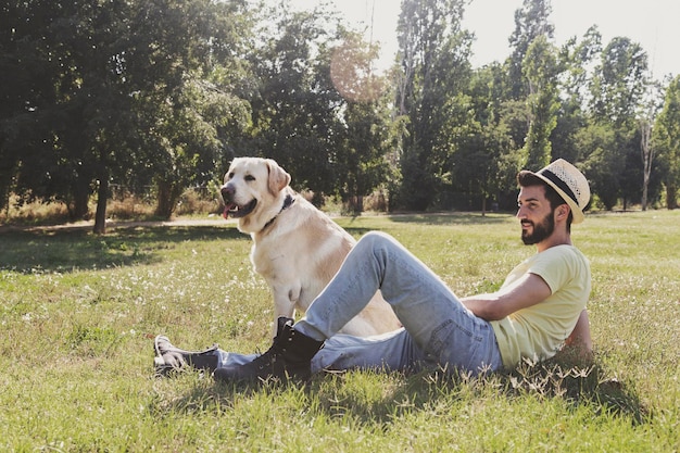 Seitliche Sicht von einem mittleren erwachsenen Mann mit einem Hund, der sich auf einem grasbewachsenen Feld gegen Bäume im Park entspannt