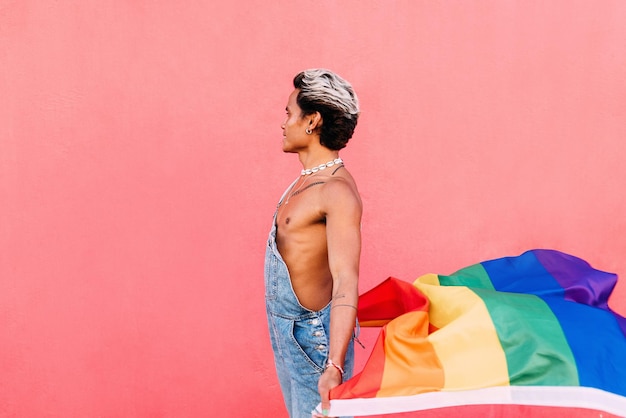 Seitliche Sicht von einem schwulen Mann, der eine Regenbogenflagge gegen einen rosa Hintergrund hält