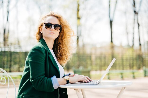 Seitliches Porträt einer süßen blonden Frau in eleganter Kleidung, die etwas auf ihrem Laptop tippt und mit nachdenklichem Ausdruck zur Seite schaut und von ihren Zukunftsplänen träumt, isoliert über dem Naturhintergrund