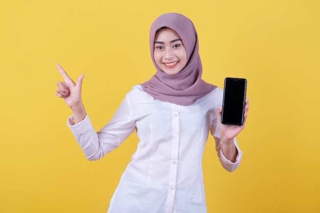 Seja esperto, compre este dispositivo, retrato de uma jovem mulher asiática rindo alto, usando um hijab apontando para cima, mostrando a tela do dispositivo