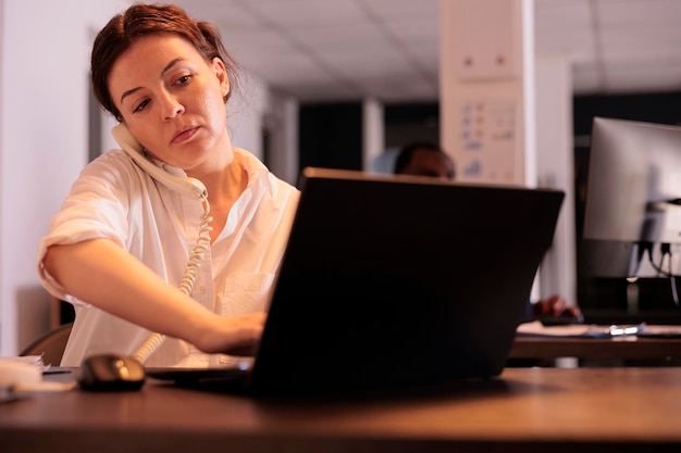 Sekretärin spricht mit Kollegin am Festnetztelefon, tippt auf Laptop, Mitarbeiter beantwortet Teamleiteranruf. Unternehmensangestellter, der nachts im Coworking Space telefoniert