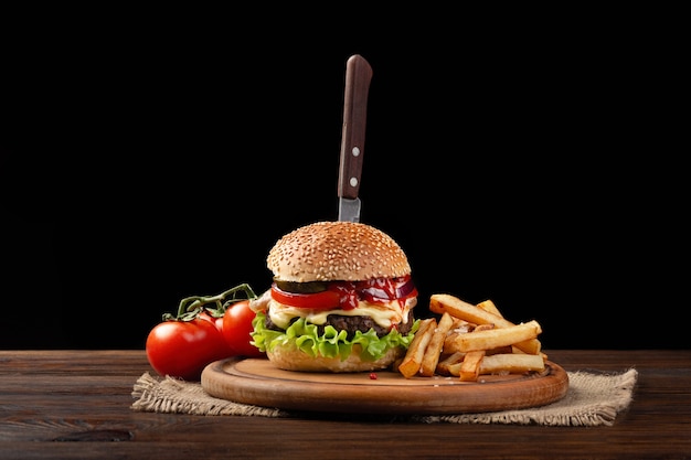 Selbst gemachte Hamburgernahaufnahme mit Rindfleisch, Tomate, Kopfsalat, Käse und Pommes-Frites auf Schneidebrett. Im Burger steckte ein Messer