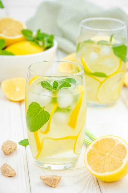 Selbst gemachte Limonade mit Zitronenscheiben, Minze und braunem Zucker in einem Glas mit Eis auf einer weißen Holzoberfläche