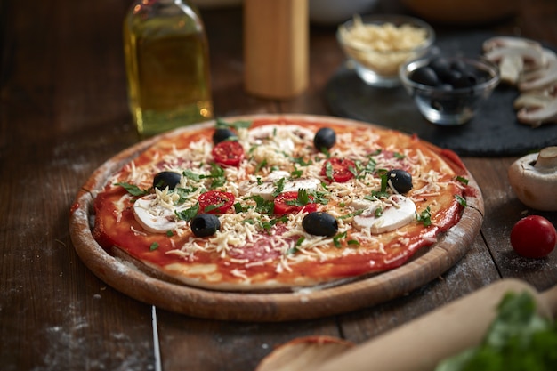 Selbst gemachte Pizza auf hölzernem Brett, mit Tomaten und Salami, Pilze, italienische Art auf altem Holztisch