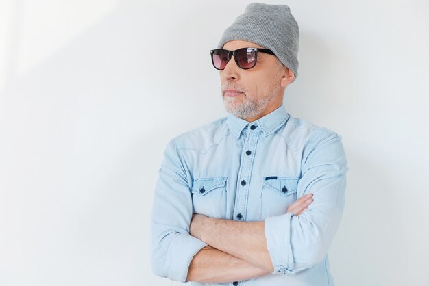 Foto selbstbewusst in seinem stil. selbstbewusster älterer mann mit grauem haar in sonnenbrille und funky hut, der wegschaut und die arme verschränkt hält, während er vor weißem hintergrund steht