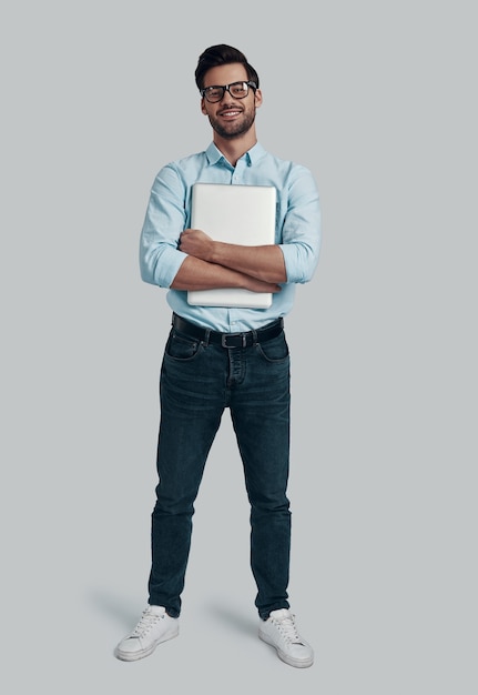 Selbstbewusst und erfolgreich. In voller Länge des jungen Mannes, der Laptop trägt und die Kamera ansieht, während er vor grauem Hintergrund steht