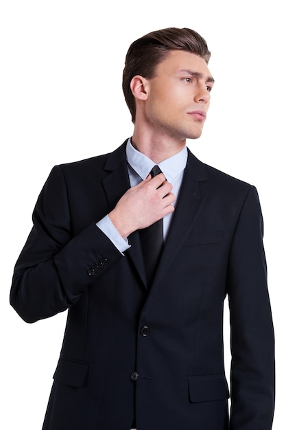 Selbstbewusst und erfolgreich. Selbstbewusster junger Mann in formeller Kleidung, der seine Krawatte anpasst und wegschaut, während er isoliert auf weiß steht