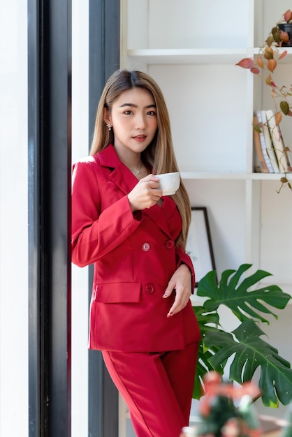 Selbstbewusste asiatische Geschäftsfrau in einem roten Anzug, die Kaffee im Büro hält und in die Kamera blickt