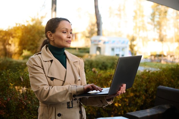 Selbstbewusste attraktive Frau mittleren Alters, freiberuflich, Geschäftsperson, die am Laptop arbeitet, im Café im Freien bei schönem herbstlichen Sonnenuntergang im Eichenhain-Park steht. Start-up, Geschäftskonzept