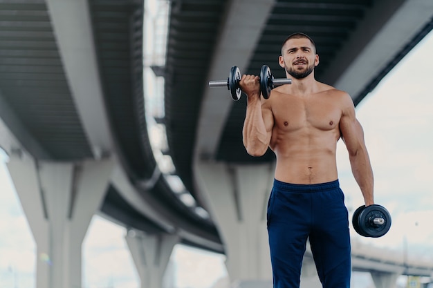 Selbstbewusste bärtige Mann Übungen mit Hanteln im Freien hat nackten Torso muskulösen hemdlosen Körper trainiert in der Nähe der Brücke macht körperliche Schwergewichtsübungen pumpt Arme Muskeln. Bodybuilding-Konzept