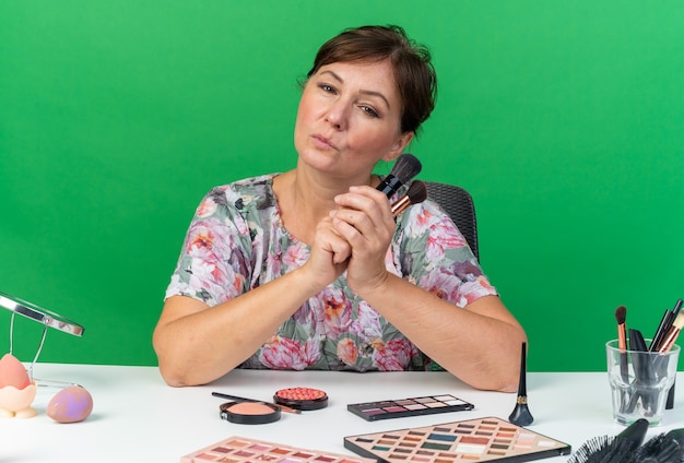 Selbstbewusste erwachsene kaukasische Frau, die am Tisch mit Make-up-Tools sitzt und Make-up-Pinsel hält