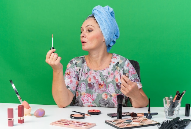 Selbstbewusste erwachsene kaukasische Frau mit eingewickeltem Haar in Handtuch, die am Tisch mit Make-up-Tools sitzt und Lipgloss hält und betrachtet