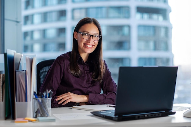 Selbstbewusste, glückliche Geschäftsfrau lächelt in die Kamera, während sie an ihrem Laptop in einem modernen Büro arbeitet, sitzt am Arbeitsplatz und hat freien Raum