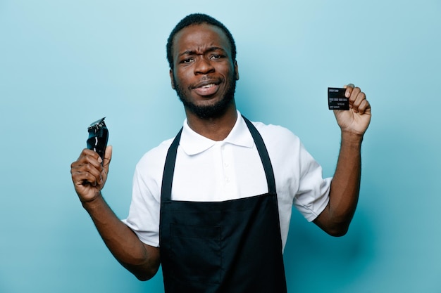 Selbstbewusste Haltekarte mit Haarschneidemaschine junger afrikanisch-amerikanischer Friseur in Uniform isoliert auf blauem Hintergrund