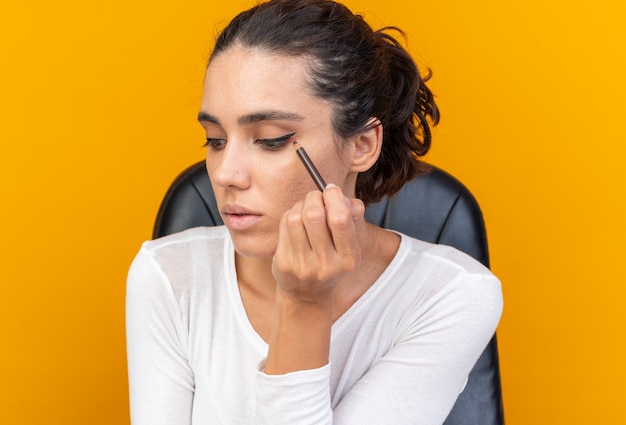 Selbstbewusste hübsche kaukasische Frau, die am Tisch mit Make-up-Tools sitzt, die Eyeliner aufträgt und die Seite isoliert auf oranger Wand mit Kopierraum betrachtet