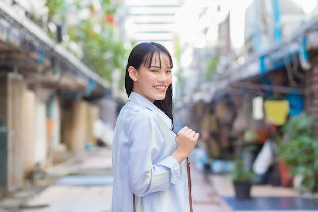 Selbstbewusste junge asiatische Frau, die ein blaues Hemd und eine Tasche trägt, lächelt glücklich, als sie zur Arbeit pendelt.