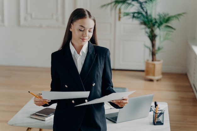 Selbstbewusste junge Geschäftsfrau in elegantem, klassischem Outfit, die Dokumente anschaut und sich mit guten Projektergebnissen glücklich fühlt, lächelnde Direktorin im modernen Büro steht und den Arbeitstag genießt enjoying